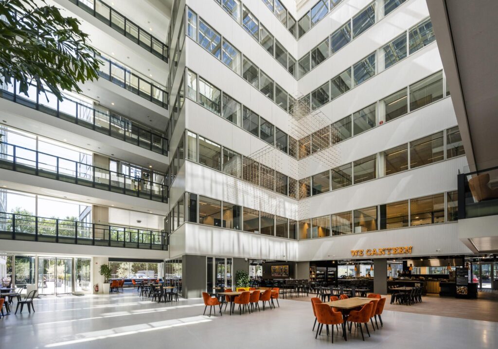 Een moderne, lichte kantoorlobby aan de Thomas R. Malthusstraat met open zitruimte en de eetgelegenheid "We Canteen".